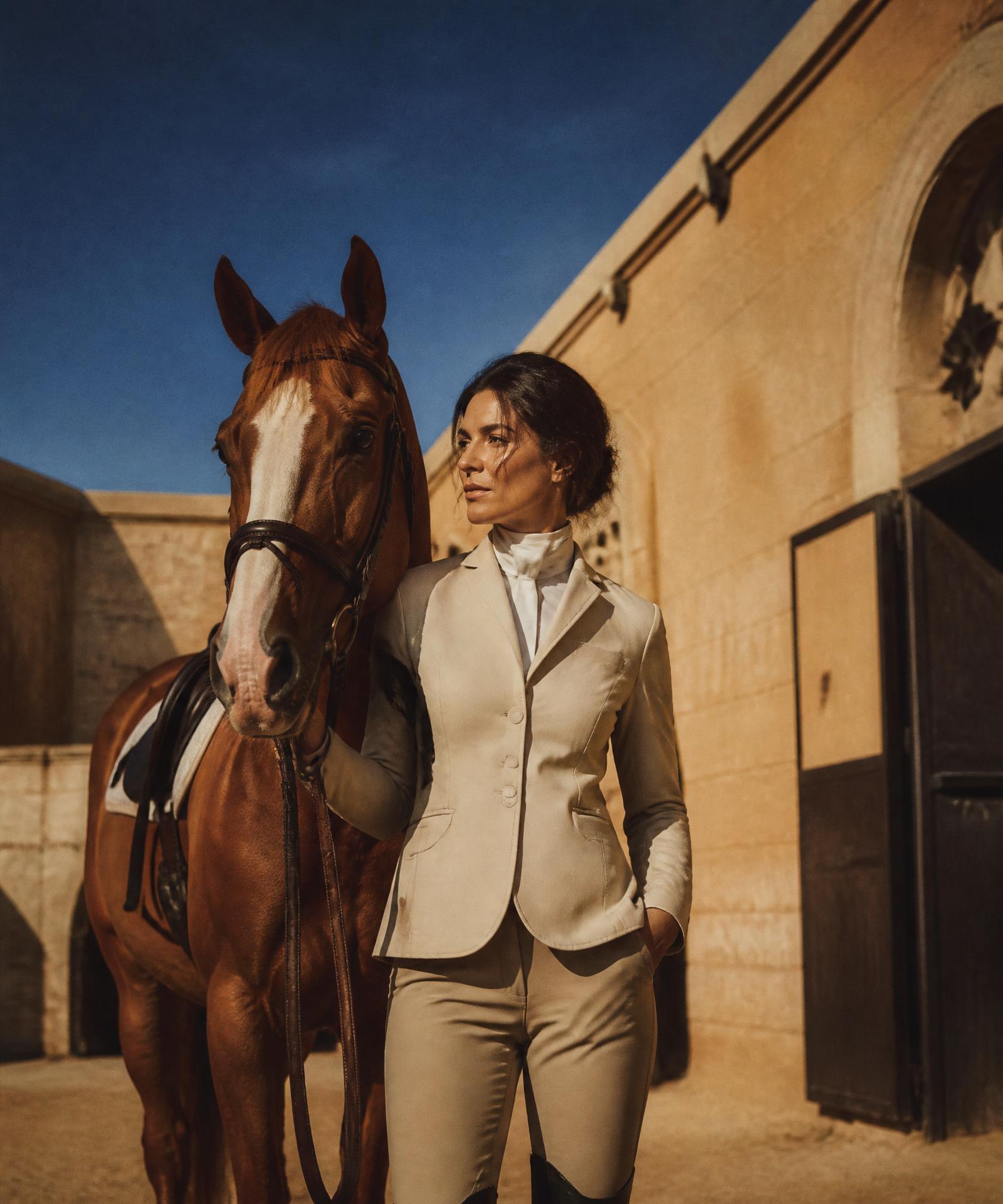 A rider in cream tailoring beside her chestnut horse in a sun-drenched sandstone courtyard